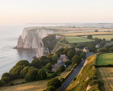 Route côtière en Normandie longeant de hautes falaises, avec un village de charme et une campagne verdoyante baignés d’une lumière douce.