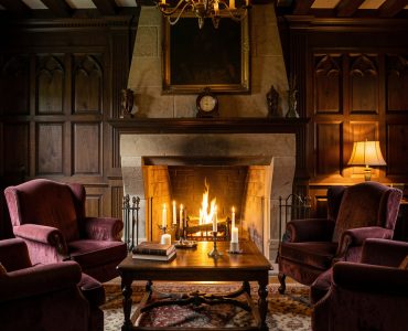 Intérieur chaleureux d’un ancien manoir normand, avec boiseries sombres, fauteuils profonds, cheminée en pierre et bougies diffusant une lumière douce dans une ambiance gothique élégante.
